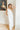 Full body view of model wearing the Go With The Flow Jumpsuit which features off white fabric, an elastic waistband, a round neckline, an upper overlay, side pockets, off white lining, a button back closure, and flared pant legs.