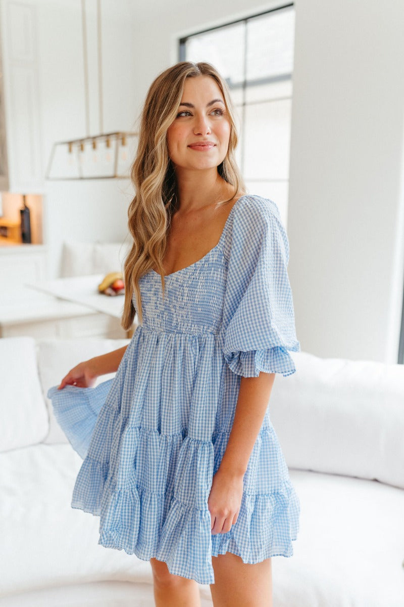 Front view of model wearing the Wanderlust Dreams Dress which features a blue and white gingham pattern, three-tiered style, mini length, white lining, a smocked upper, a sweetheart neckline, and 3/4 puff sleeves with ruffle hem.