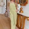 Full body view of model wearing the Step On It Jumpsuit which features olive green fabric, two front pockets, two back pockets, an elastic waistband, a square neckline, spaghetti straps, an open back with ties, and flared pant legs.