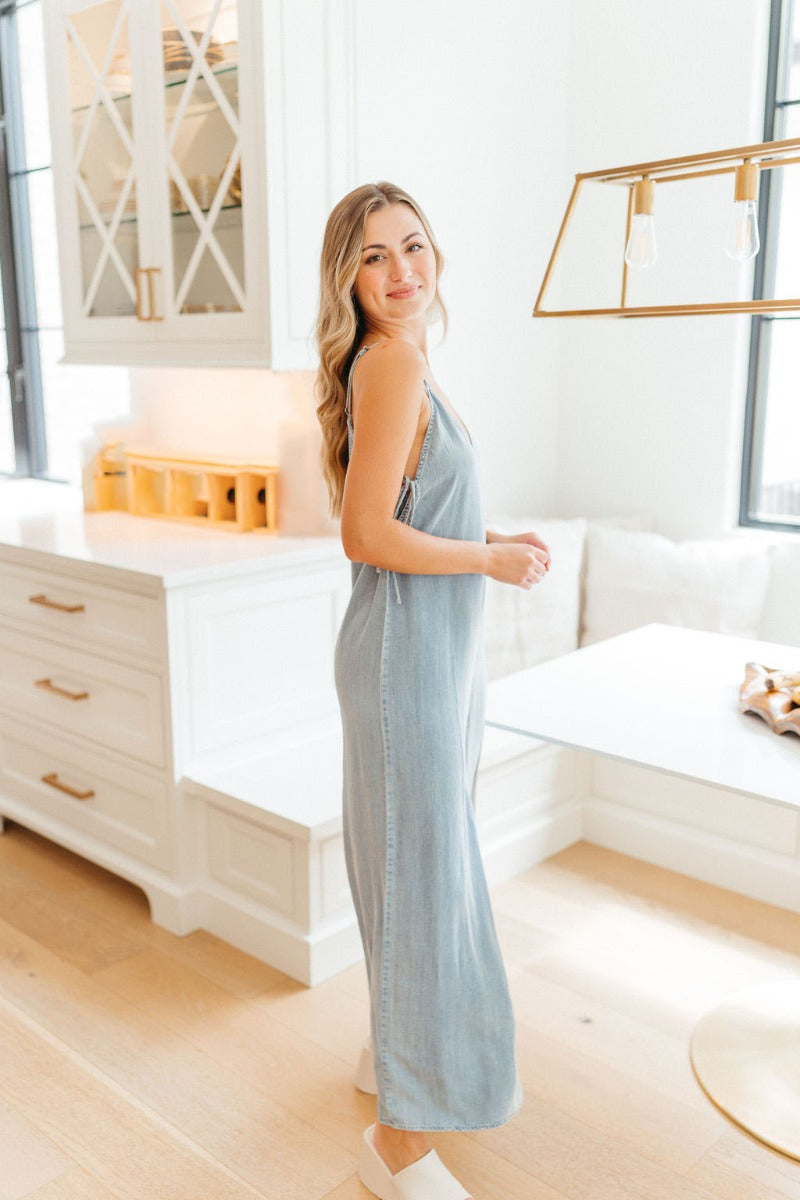 Side view of model wearing the See You Again Jumpsuit which features light denim fabric, open side ties, a v neckline, adjustable spaghetti straps, a back zipper, and wide pant legs.
