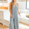 Full body view of model wearing the See You Again Jumpsuit which features light denim fabric, open side ties, a v neckline, adjustable spaghetti straps, a back zipper, and wide pant legs.