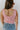 Back view of model wearing the Picking Daisies Top in Pink that has light pink crochet knit fabric with a cream daisy pattern, a cropped waist, scalloped hem, a scoop neck, and thick straps.
