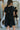 Back view of model wearing the Never Let Me Go Dress that has black cotton fabric, a mini length hem, diagonal tiered style, a round neck, a back keyhole with a tie, and short puff sleeves.