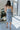 Back view of model wearing the Better This Way Dress which features light blue, dark blue and cream brocade fabric with a floral design, a midi-length hem, a front slit, a straight strapless neckline, and a monochromatic back zipper.