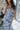Close up view of model wearing the Better This Way Dress which features light blue, dark blue and cream brocade fabric with a floral design, a midi-length hem, a front slit, a straight strapless neckline, and a monochromatic back zipper.