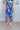 Close up view of model wearing the Meet Me At The Beach Skirt which features white fabric with a blue leaf design, white lining, midi length, a slit on the side, and a wrap waist with a tie and button closure.