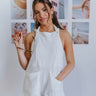 Full body view of model wearing the On Island Time Romper which features ivory fabric, textured front pockets, a halter neckline that ties around the neck, and an open back with an elastic band.
