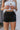 Front view of model wearing the Rolling Stones Shorts which features black denim fabric, silver stud details on the side, a front zipper and button closure, belt loops, two front pockets, two back pockets and a frayed hem.