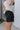 Side view of model wearing the Rolling Stones Shorts which features black denim fabric, silver stud details on the side, a front zipper and button closure, belt loops, two front pockets, two back pockets and a frayed hem.