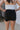 Back view of model wearing the Rolling Stones Shorts which features black denim fabric, silver stud details on the side, a front zipper and button closure, belt loops, two front pockets, two back pockets and a frayed hem.