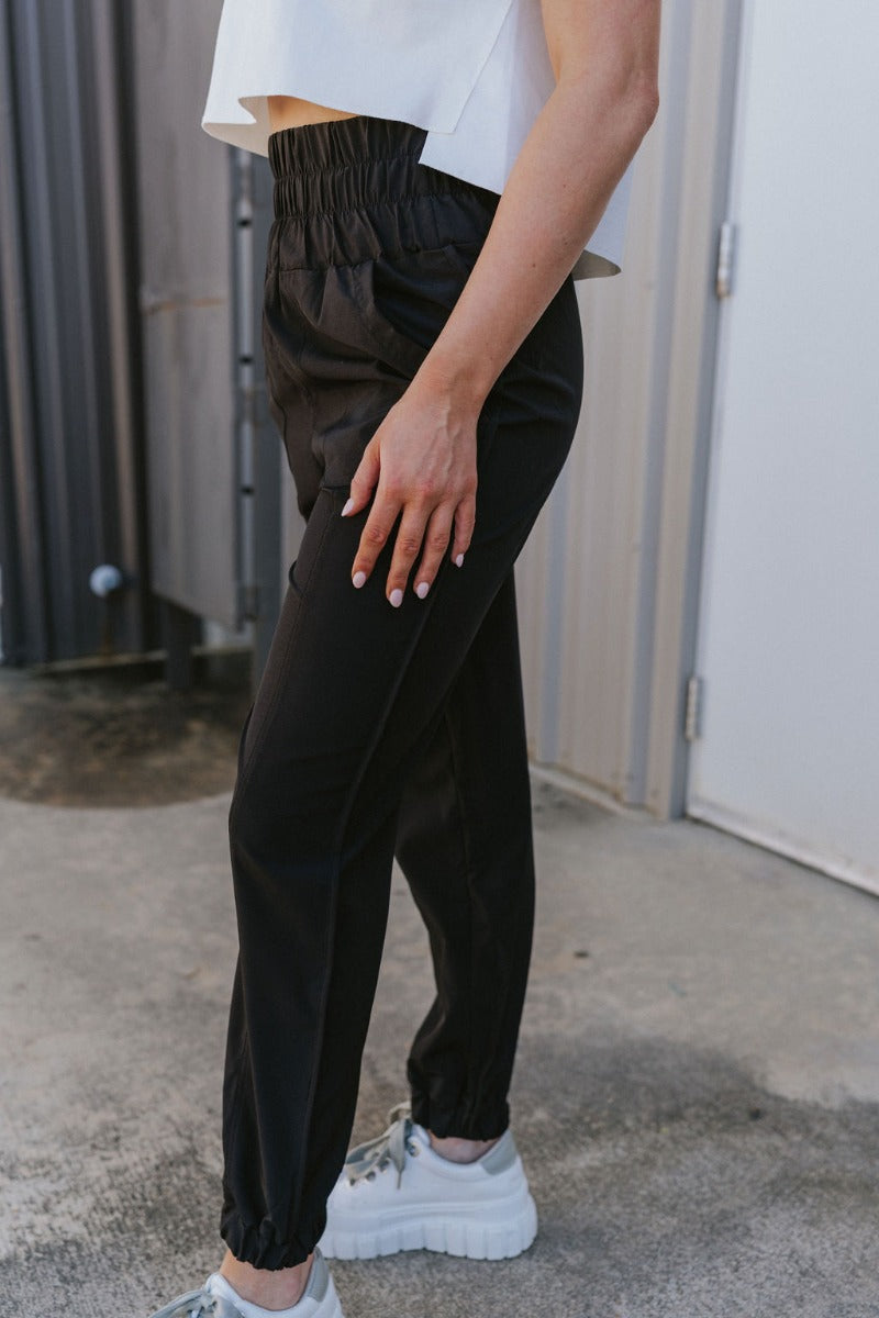 Side view of model wearing the Whatever It Takes Joggers which features black lightweight fabric, side pockets, a smocked high-rise waisted band, and jogger legs with elastic ankles.