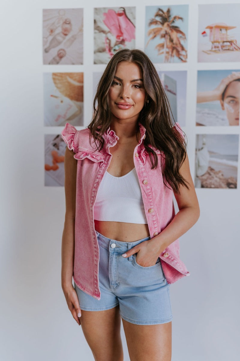 Front view of model wearing the Hidden Romance Top which features pink washed denim fabric, light tortoise buttons, a high neckline, ruffle details, and a sleeveless design.