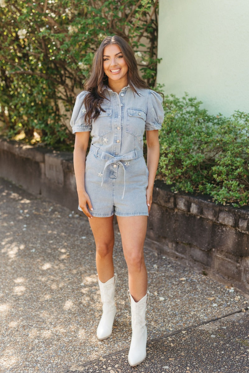 Full front view of model wearing the She's A Star Denim Romper that has light denim fabric, pockets on each side, silver buttons, silver sequin star patches, a collared neckline, short puff sleeves, and denim tie belt around the waist