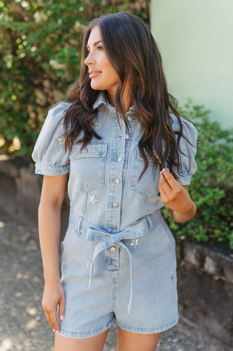 Front view of model wearing the She's A Star Denim Romper that has light denim fabric, pockets on each side, silver buttons, silver sequin star patches, a collared neckline, short puff sleeves, and denim tie belt around the waist