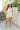 Full body front view of model wearing the Great Escape Overalls that have cream denim fabric, pockets, silver buttons, belt loops, a square neckline, adjustable straps, and distressed details