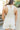 Back view of model wearing the Great Escape Overalls that have cream denim fabric, pockets, silver buttons, belt loops, a square neckline, adjustable straps, and distressed details