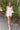 Full body front view of model wearing the Happiest With You Romper that has white denim fabric, a scooped skirt design with ruched detailing on the side, white shorts lining, a strapless sweetheart neck, and a back zipper with hook closure