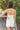 Back view of model wearing the Happiest With You Romper that has white denim fabric, a scooped skirt design with ruched detailing on the side, white shorts lining, a strapless sweetheart neck, and a back zipper with hook closure
