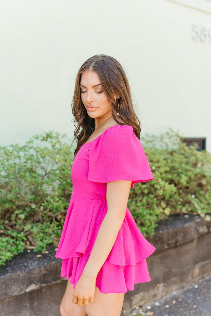 Side view of model wearing the Wishful Thinking Romper which features fuchsia fabric, a ruffle-tiered skirt, fuchsia shorts lining, an elastic waistband, a sweetheart neckline and short puff sleeves.