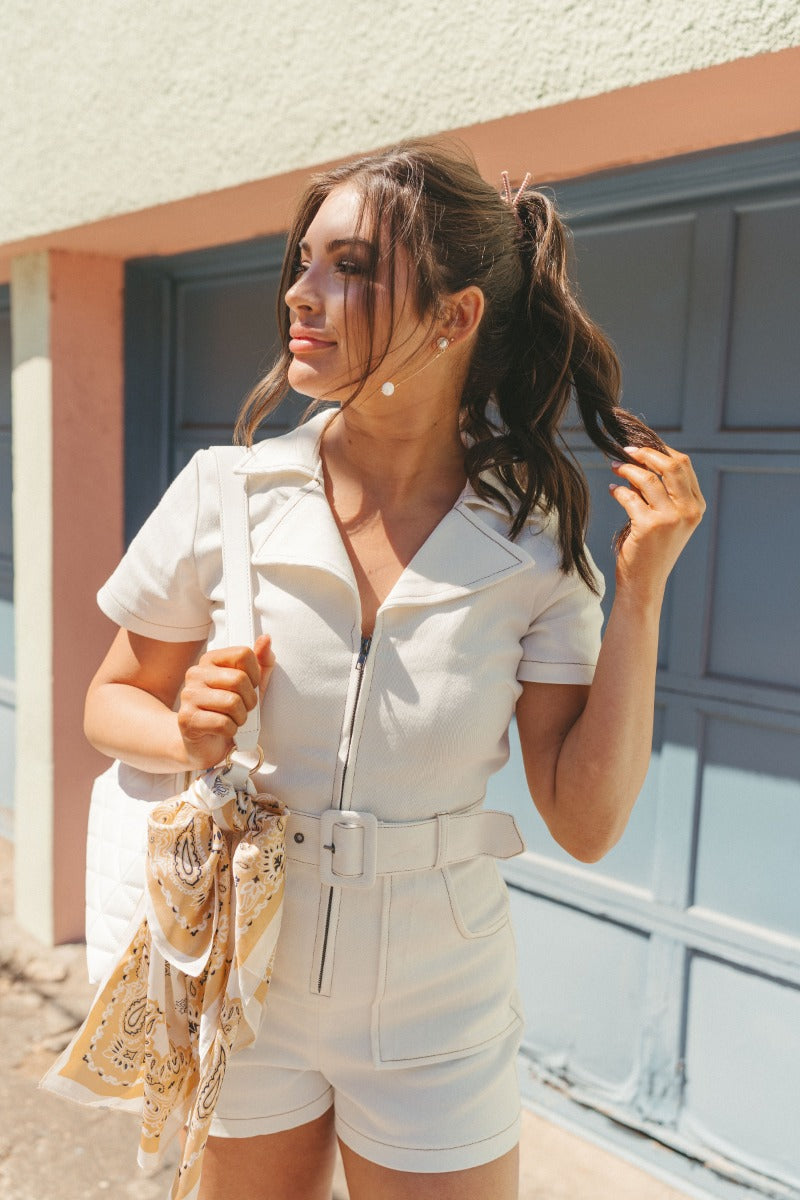 Front view of model wearing the Dakota Denim Romper in Off White which features white denim fabric, front pockets, a hidden front zipper, a collared neckline, a monochromatic belt at the waistline, and short sleeves.