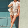 Full body view of model wearing the Dakota Denim Romper in Off White which features white denim fabric, front pockets, a hidden front zipper, a collared neckline, a monochromatic belt at the waistline, and short sleeves.