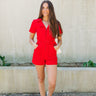 Full body view of model wearing the Dakota Denim Romper in Red which features red denim fabric, front pockets, a hidden front zipper, a collared neckline, a monochromatic belt at the waistline, and short sleeves.