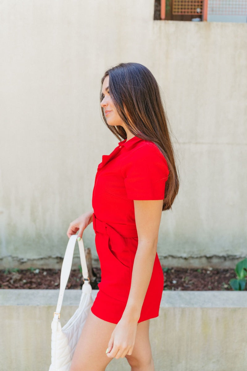 Side view of model wearing the Dakota Denim Romper in Red which features red denim fabric, front pockets, a hidden front zipper, a collared neckline, a monochromatic belt at the waistline, and short sleeves.