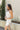 Side view of model wearing the Starlight Rhinestone Fringe Dress in White which features white fabric, a straight neckline, a sleeveless design with thin adjustable straps, rhinestone fringe detailing at the bottom side hem, criss cross straps in the back