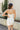 Back view of model wearing the Starlight Rhinestone Fringe Dress in White which features white fabric, a straight neckline, a sleeveless design with thin adjustable straps, rhinestone fringe detailing at the bottom side hem, criss cross straps in the back