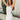 Side view of model wearing the Starlight Rhinestone Fringe Dress in White which features white fabric, a straight neckline, a sleeveless design with thin adjustable straps, rhinestone fringe detailing at the bottom side hem, criss cross straps in the back