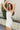 Side view of model wearing the Starlight Rhinestone Fringe Dress in White which features white fabric, a straight neckline, a sleeveless design with thin adjustable straps, rhinestone fringe detailing at the bottom side hem, criss cross straps in the back