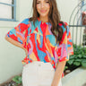 Front view of model wearing the Pop Of Color Blouse which features red fabric with a blue, orange, green, purple, pink and yellow print, a round neckline, a back keyhole with a button closure, and short flare sleeves.