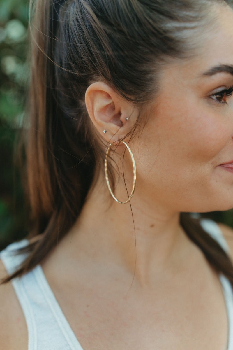 Side view of model wearing the On Your Own Hoop Earrings in Gold that feature large closed gold hoops with a hammered design.