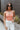 Front view of model wearing the Chasing Summer Tank in Peach which features peach knit fabric, a cropped waist, a halter neckline, an open back and a sleeveless design.