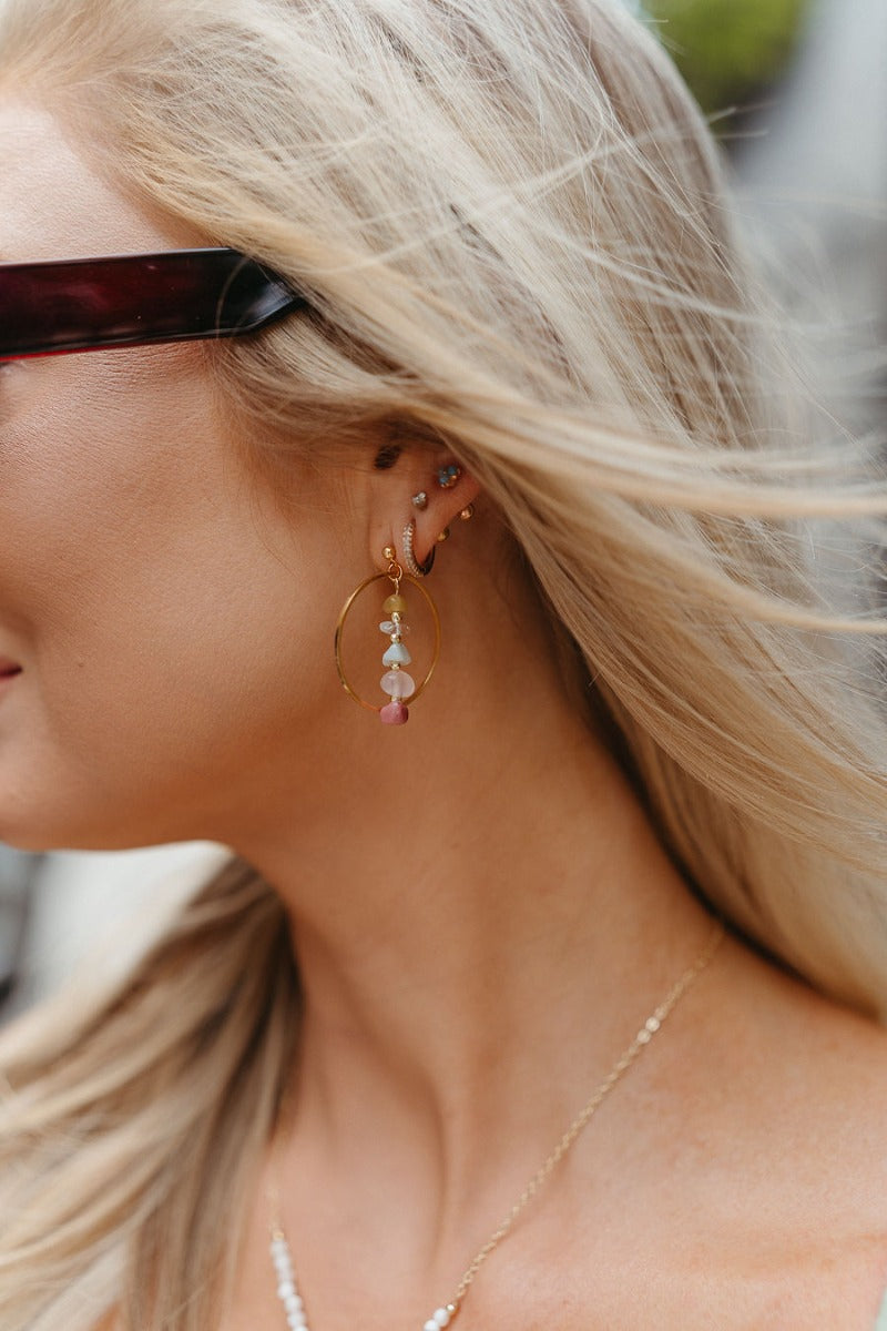Side view of model wearing the Take Me Away Multi Earring which features gold open circle attached to a gold chain link with purple, light purple, aqua, clear and green beads.
