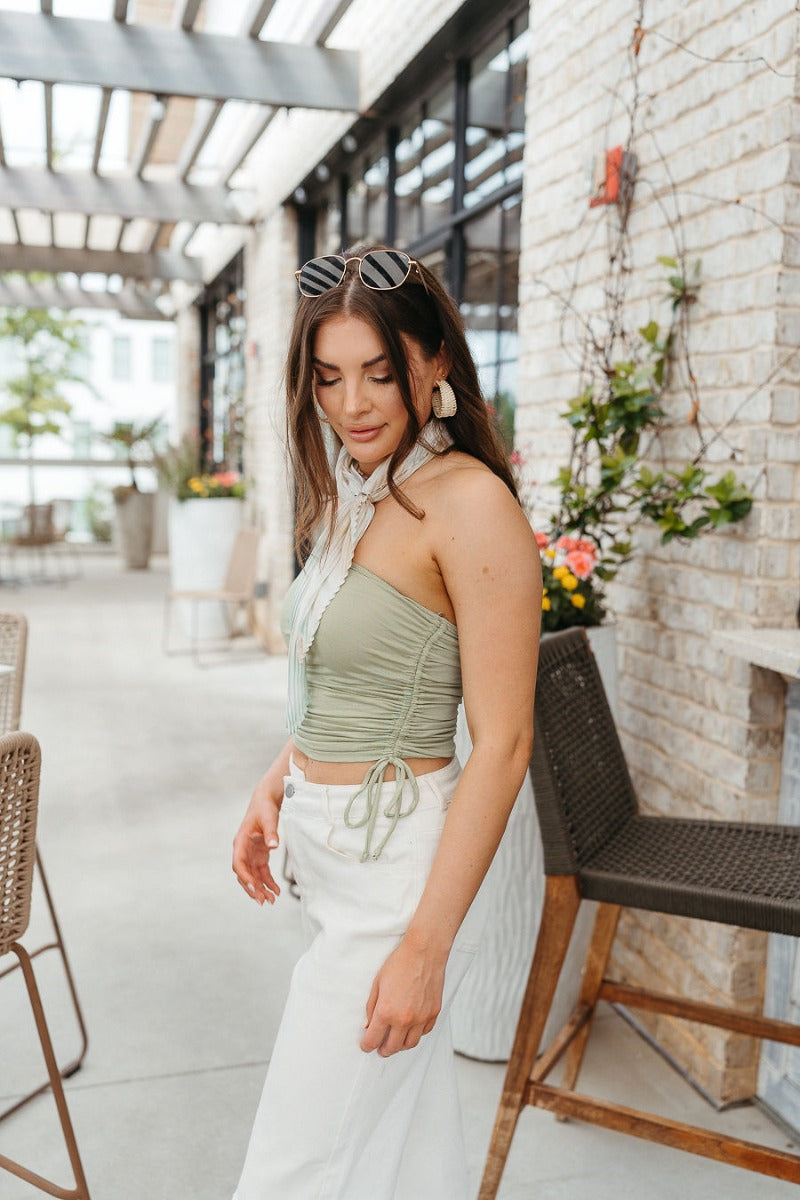 Side view of model wearing the Dare To Be Different Crop Top which features light sage knit fabric, ruched side details with ties, a cropped waist and a strapless neckline.