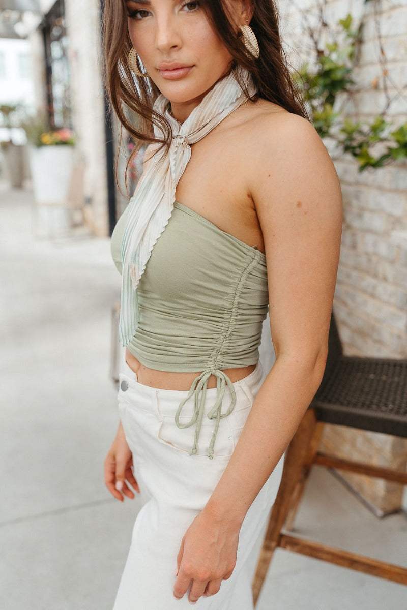 Side view of model wearing the Dare To Be Different Crop Top which features light sage knit fabric, ruched side details with ties, a cropped waist and a strapless neckline.