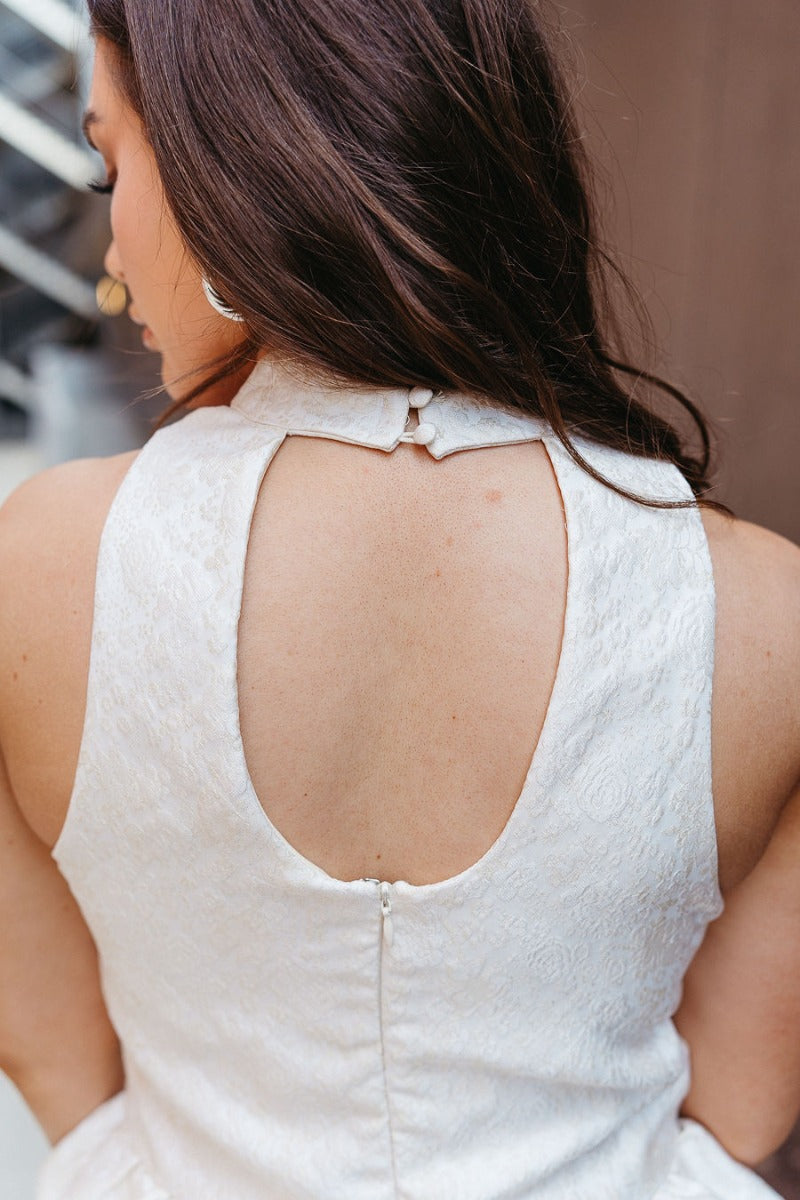 Close up back view of model wearing the Full of Class Top which features white fabric with a cream embroidered rose pattern, a ruffle tiered hem, a high neckline, a keyhole back with buttons and a hook closure, and a sleeveless design.