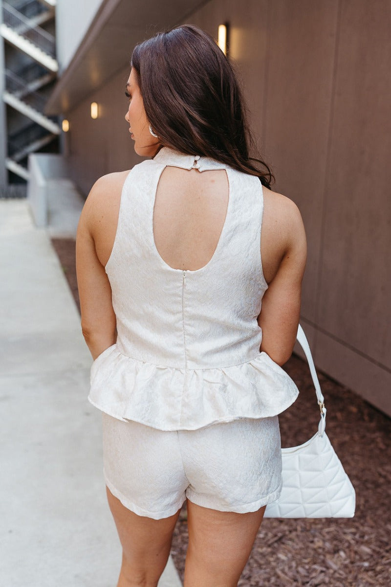 Back view of model wearing the Full of Class Shorts which features white fabric with a cream embroidered rose pattern, cream shorts lining, front pockets, and a monochromatic side zipper with a hook closure.