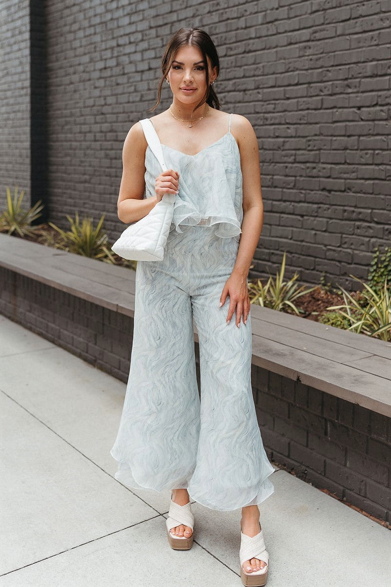 Full body front view of model wearing the Sea Breeze Pants that have white and sage sheer fabric with a marbled pattern, flared legs with a ruffle tier hem, and a  side zipper.