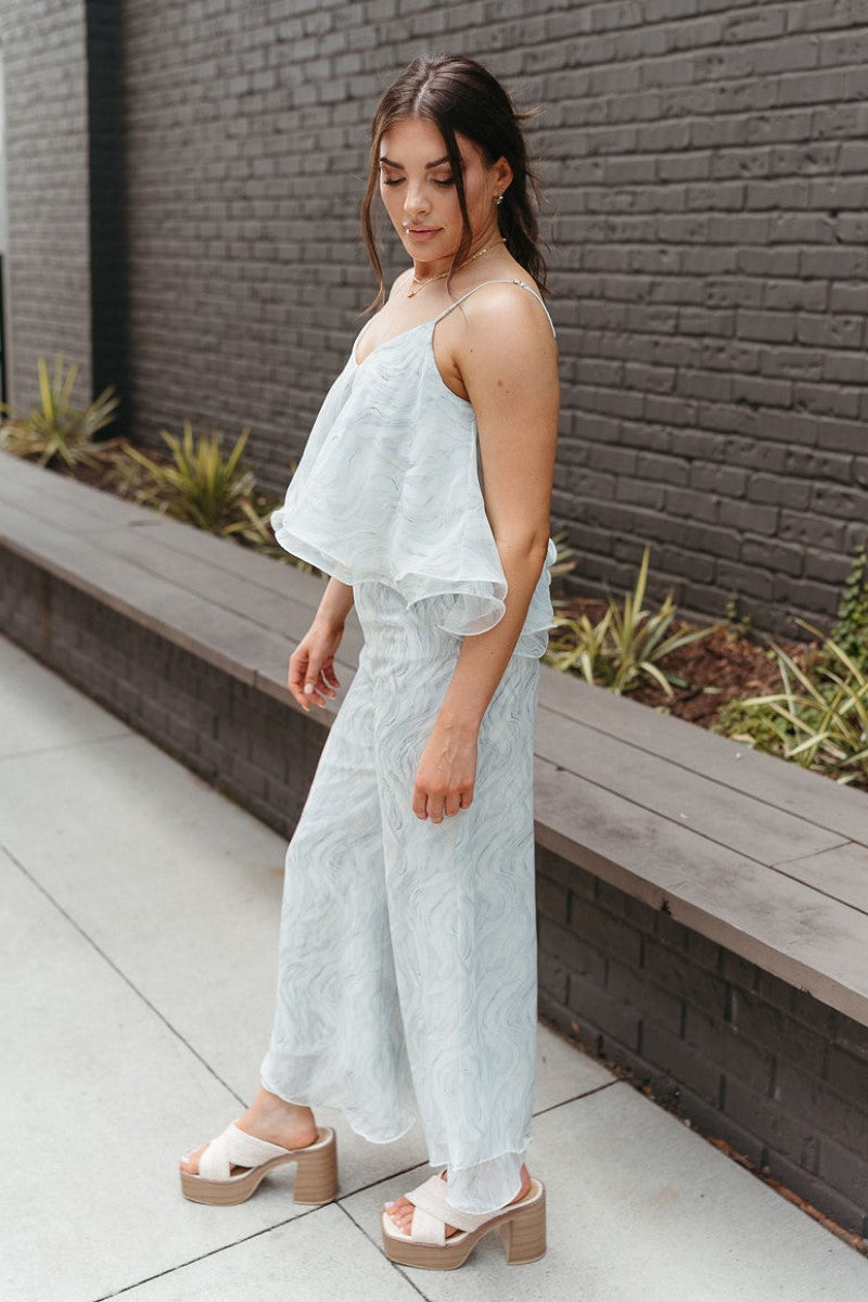 Full body side view of model wearing the Sea Breeze Pants that have white and sage sheer fabric with a marbled pattern, flared legs with a ruffle tier hem, and a  side zipper.