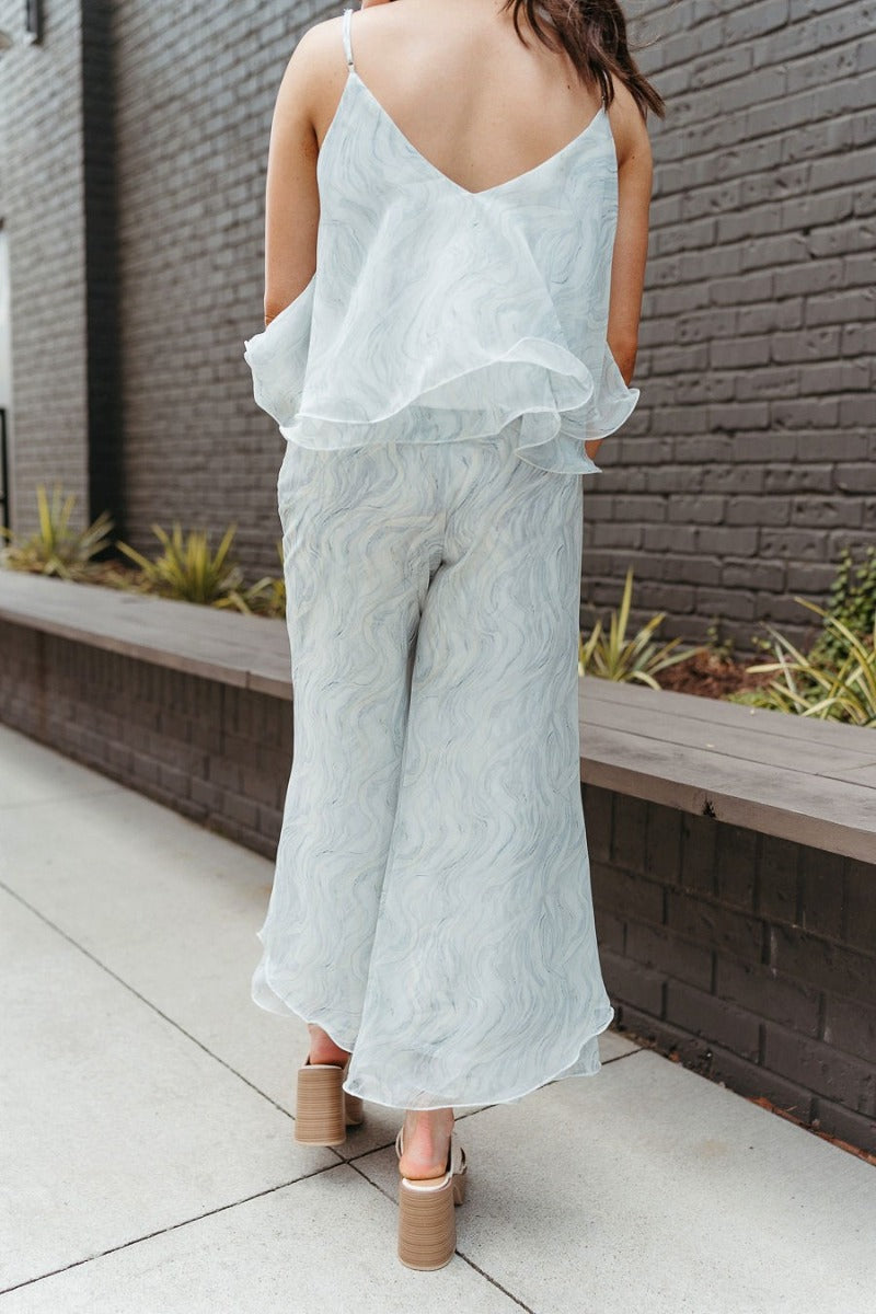 Back view of model wearing the Sea Breeze Pants that have white and sage sheer fabric with a marbled pattern, flared legs with a ruffle tier hem, and a  side zipper.