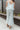 Back view of model wearing the Sea Breeze Pants that have white and sage sheer fabric with a marbled pattern, flared legs with a ruffle tier hem, and a  side zipper.