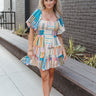Full body front view of model wearing the Mosaic Views Dress that has a box design with vertical and horizontal stripes, a ruffle hem, a smocked upper, a square neck, and short puff sleeves.