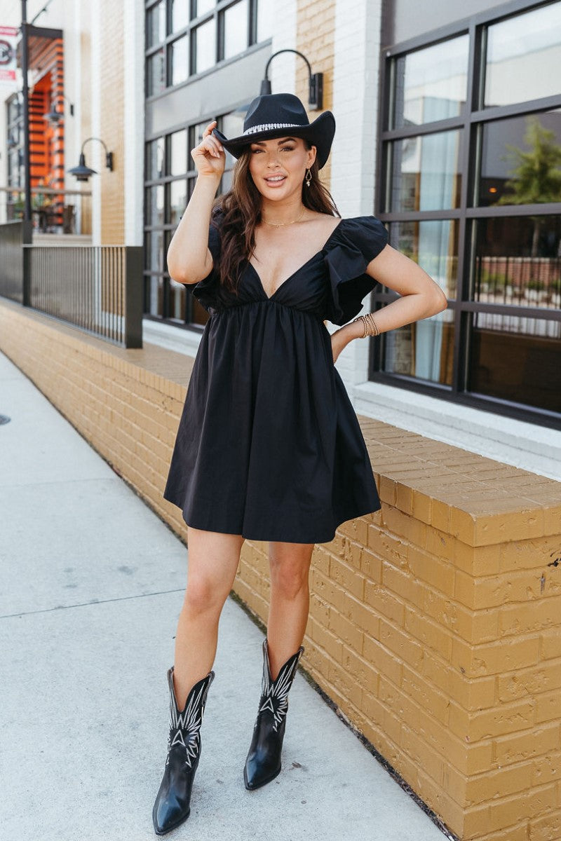 Full body front view of model wearing the Heart Breaker Dress In Black that has black fabric, a mini-length hem, a baby doll style, a v neck, elastic straps with ruffles, and an open back with a bow.
