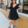 Full body front view of model wearing the Heart Breaker Dress In Black that has black fabric, a mini-length hem, a baby doll style, a v neck, elastic straps with ruffles, and an open back with a bow.