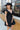 Frontal view of model wearing the Heart Breaker Dress In Black that has black fabric, a mini-length hem, a baby doll style, a v neck, elastic straps with ruffles, and an open back with a bow.