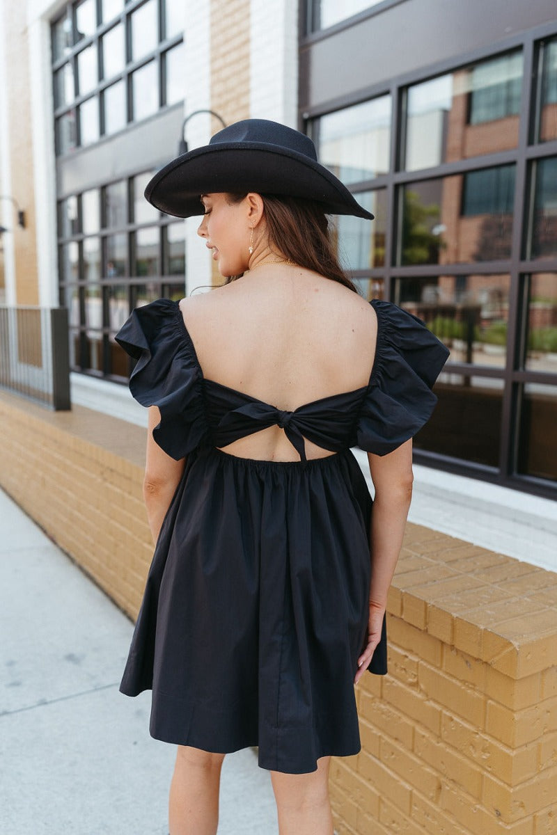 Back view of model wearing the Heart Breaker Dress In Black that has black fabric, a mini-length hem, a baby doll style, a v neck, elastic straps with ruffles, and an open back with a bow.