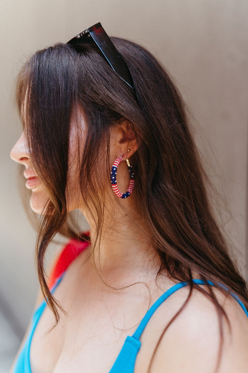 Side view of model wearing the Proud To Be Earring which features red and white stripe beads with navy blue beads and white polka dots wrapped on closed gold hoops.