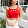 Front view of model wearing the Party In The USA Tank which features red knit fabric, a cropped waist, a square neckline, adjustable straps, and "USA" stitched in white.